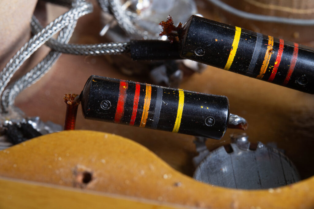 Close-up of two original "Bumblebee" capacitors inside a vintage Gibson Les Paul Special control cavity. The capacitors feature a black cylindrical body with distinctive colorful stripes (indicating capacitance and voltage) and are soldered to Centralab (CRL) potentiometers. The surrounding mahogany wood shows original factory routing, a rubber grommet for wiring, and authentic 1950s solder joints.